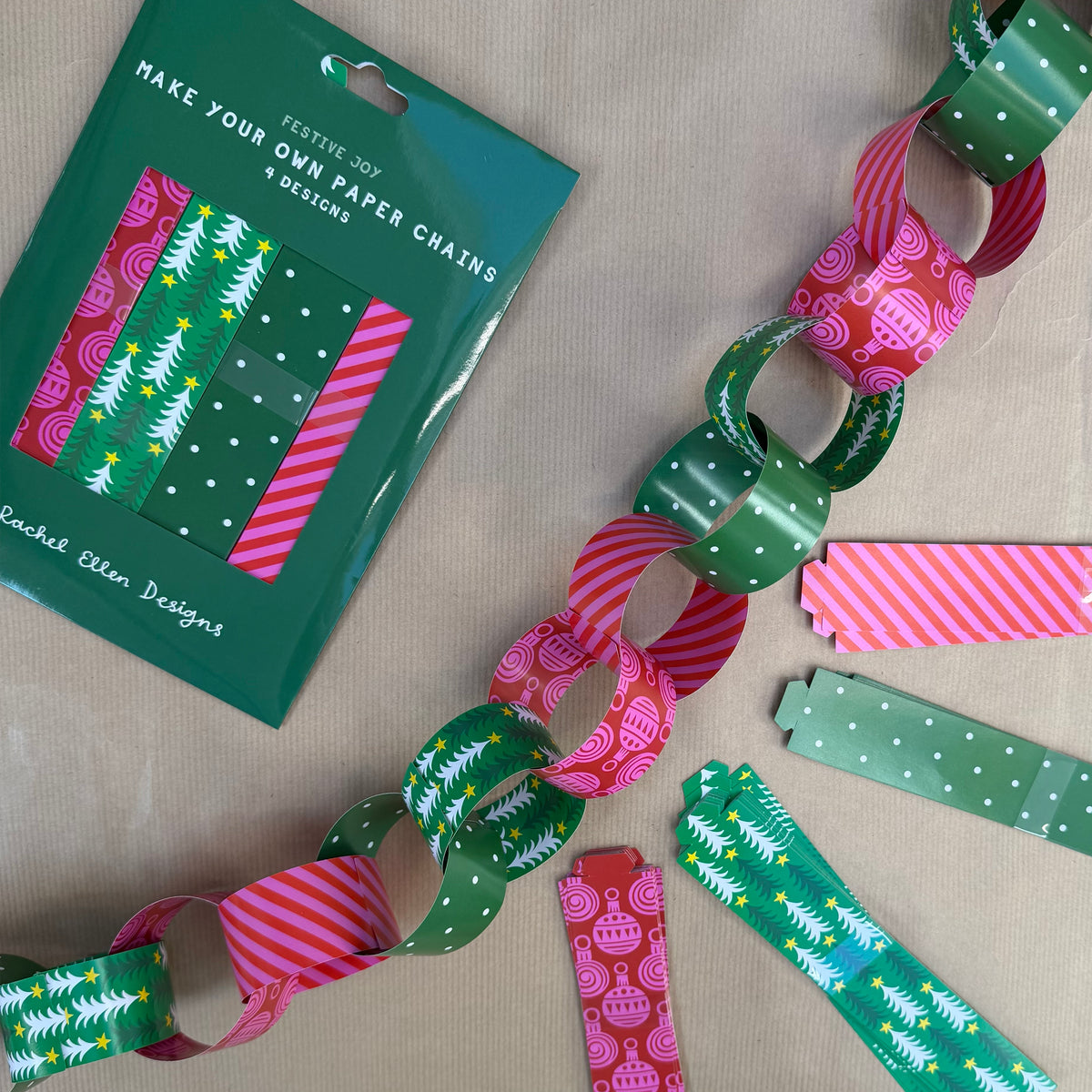 A festive paper chain garland made of green, pink, and white holiday-patterned paper strips, displayed on a neutral beige fabric alongside its green retail packaging card.